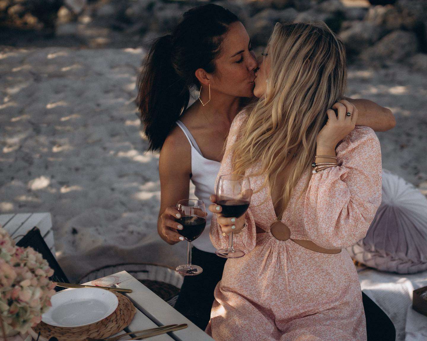 Two women seated on the beach, sharing a bottle of wine, with the sun shining and the ocean in the background.