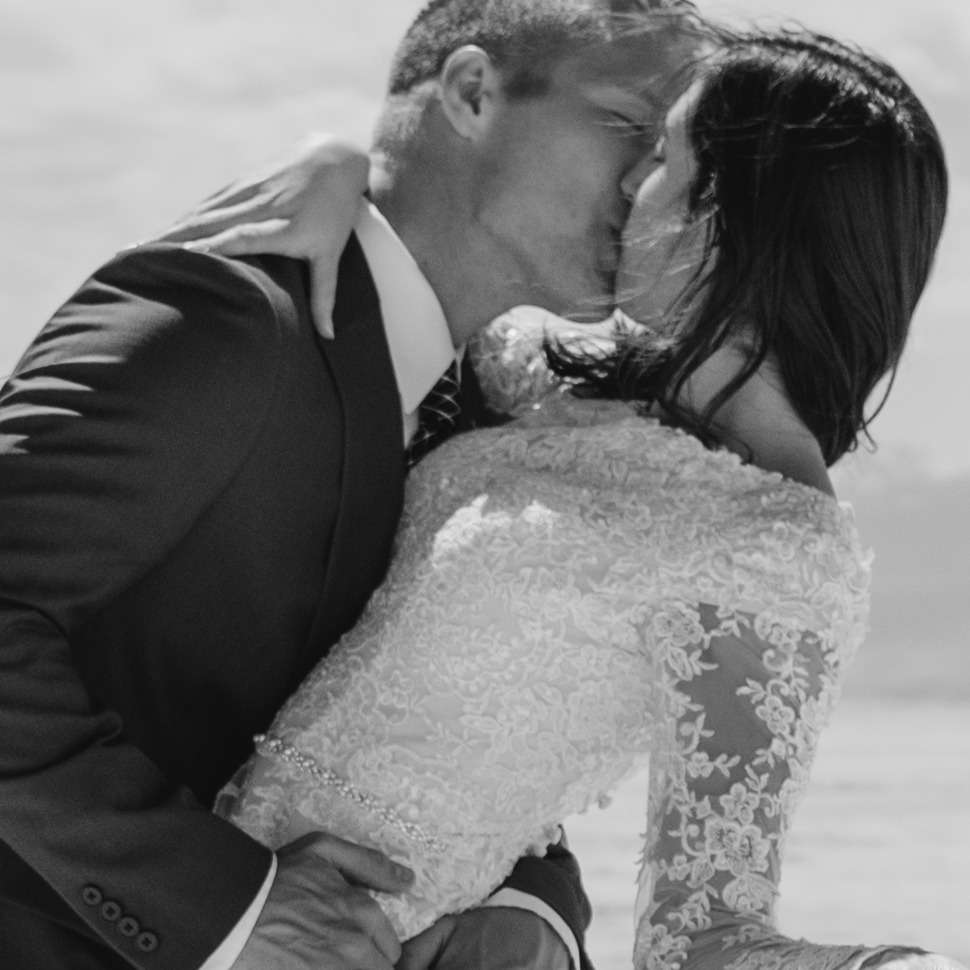 A bride and groom share a romantic kiss on a picturesque beach, with waves gently lapping at the shore in the background.